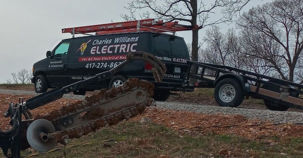The black Charles Williams Electric van with ladders on the roof rack and a utility trailer, on a rural job with a trencher boom in the foreground